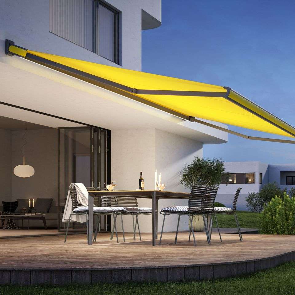 eine gelbe kassettenmarkise markilux mx 3 mit integrierter led beleuchtung erleuchtet eine holzterrasse am abend