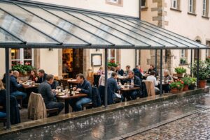 Gäste sitzen bei Regen trocken unter einem Glasdach auf einer Restaurantterrasse